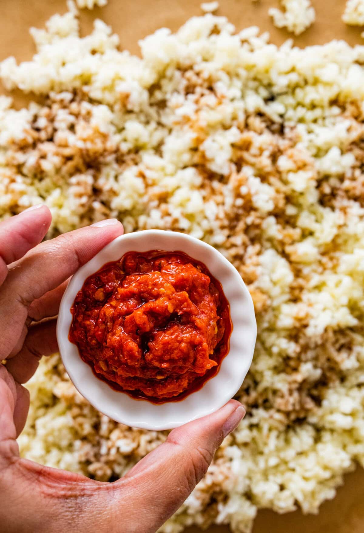 A hand holding a small bowl of red sauce over a pile of rice