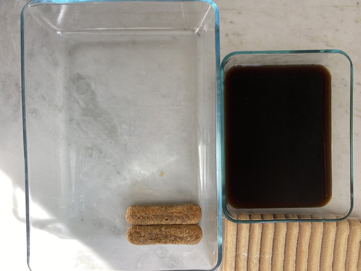 A pan with coffee mixture and a couple dipped lady fingers in the dish to make tiramisu.