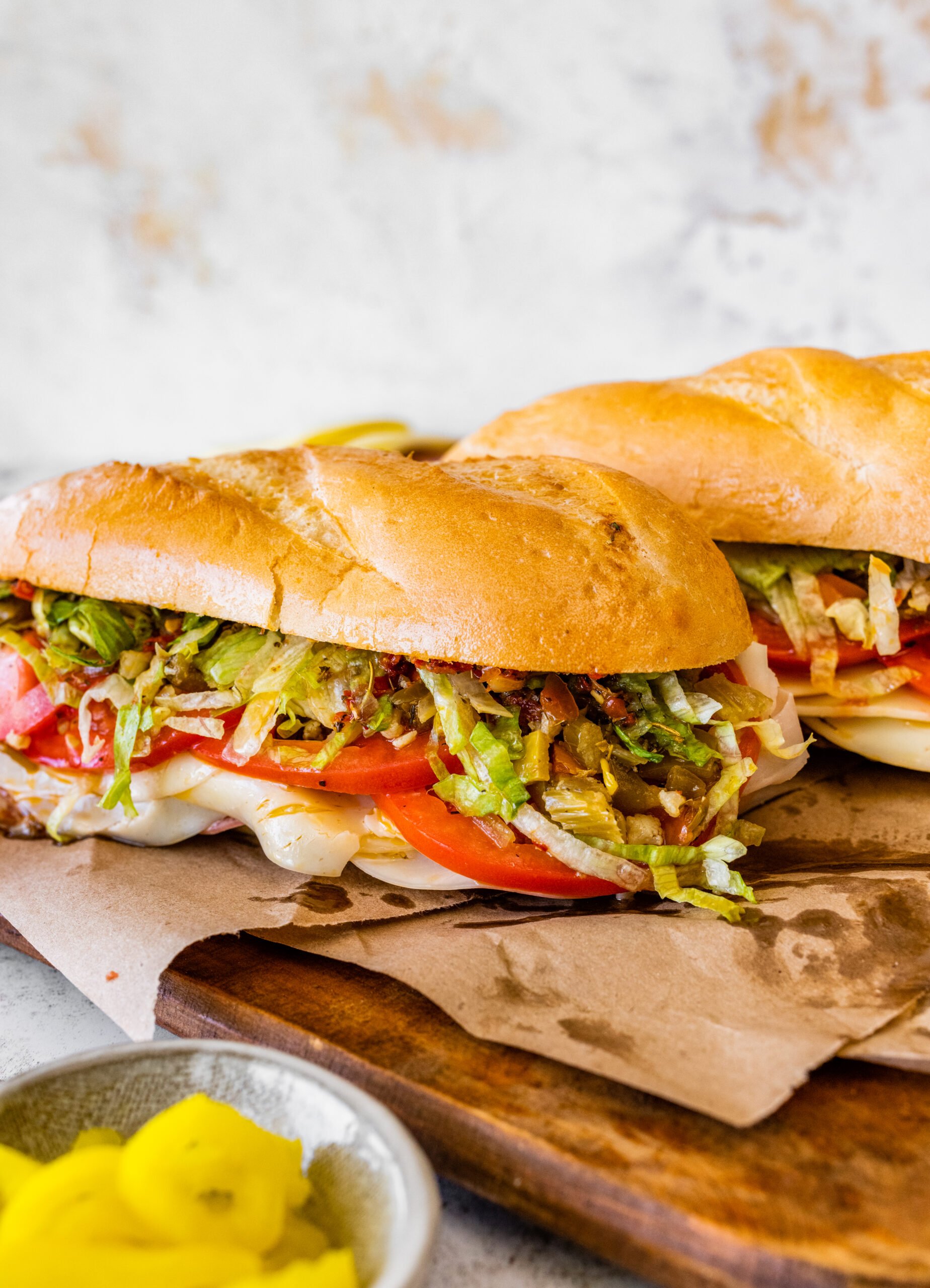Two Italian sub sandwiches on a cutting board with condiments on the side.
