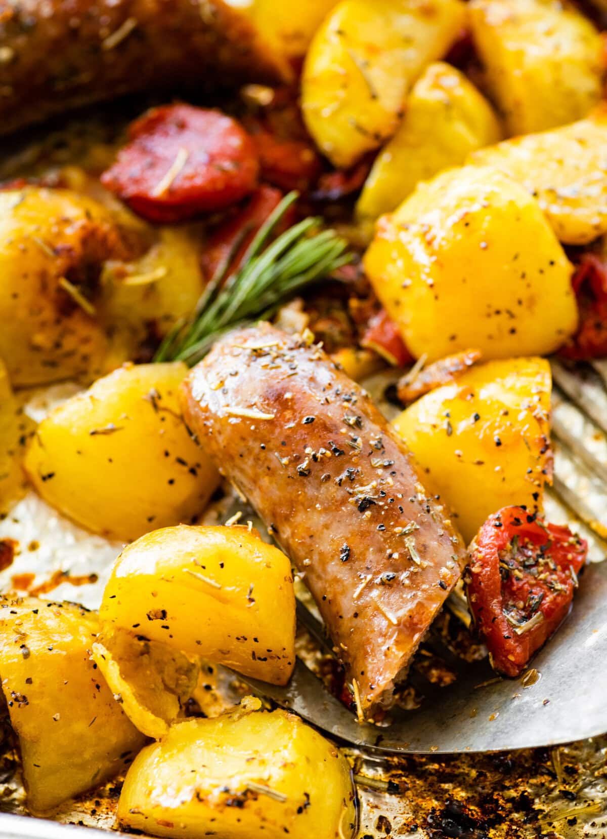 A serving spoon lifting some sausage and potatoes from the baking pan.