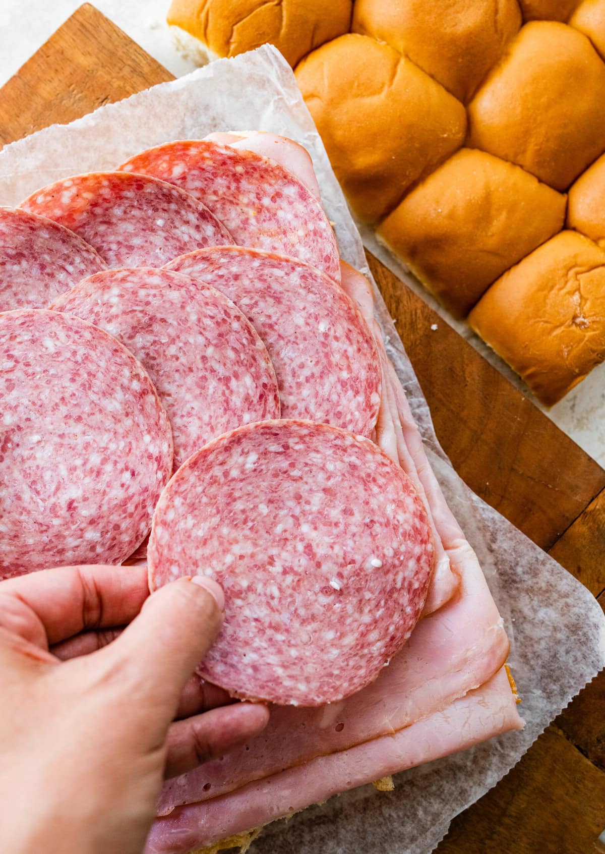A person placing a slice of ham and salami on a tray for the Italian sliders.