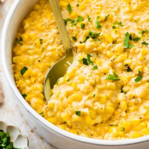creamed corn recipe in a white bowl with a little parsley on top and extra cheese. Spoon in the bowl.