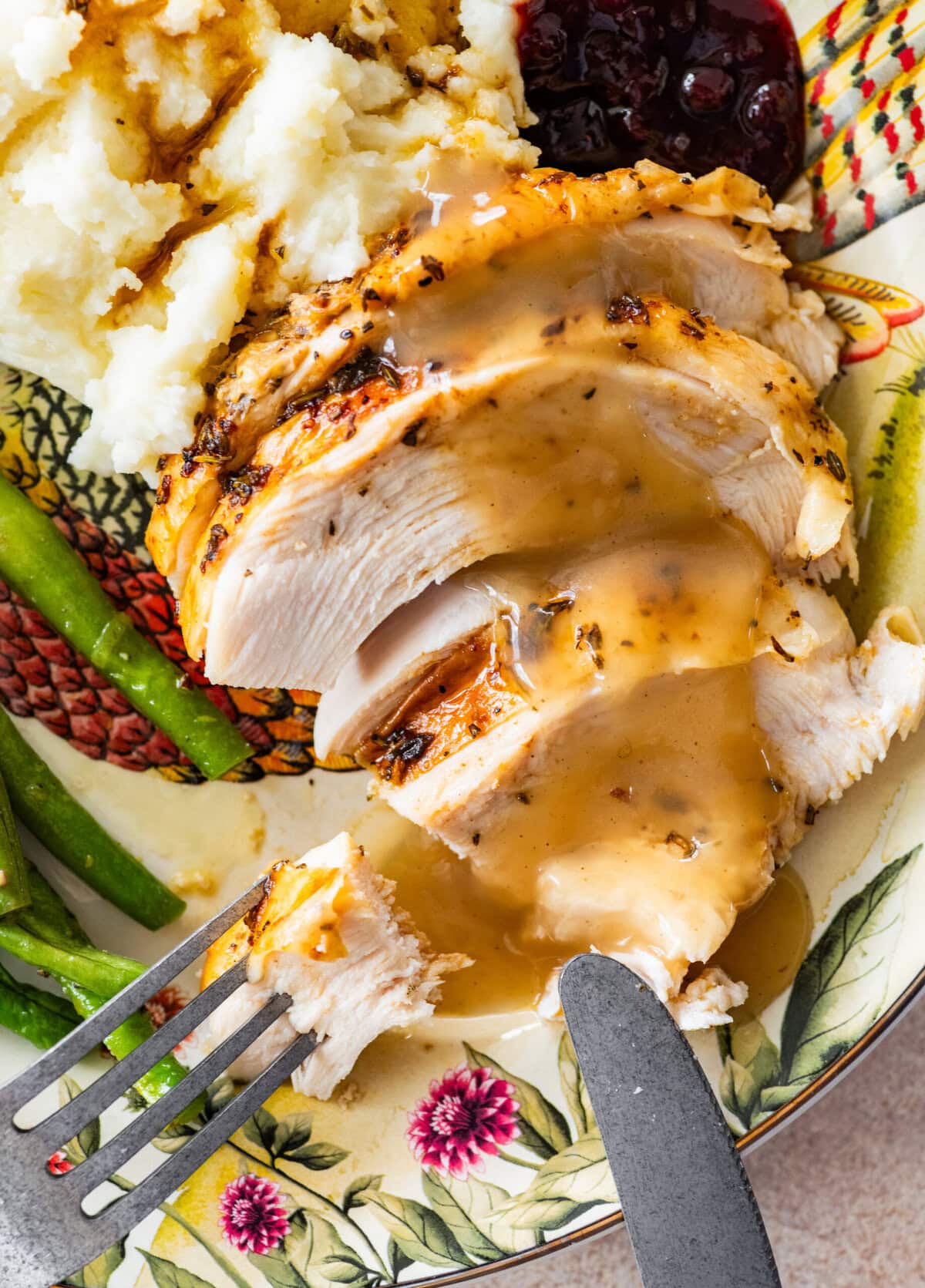 roast turkey breast slices on a plate with mashed potatoes, green beans, and gravy. Fork and knife on plate.