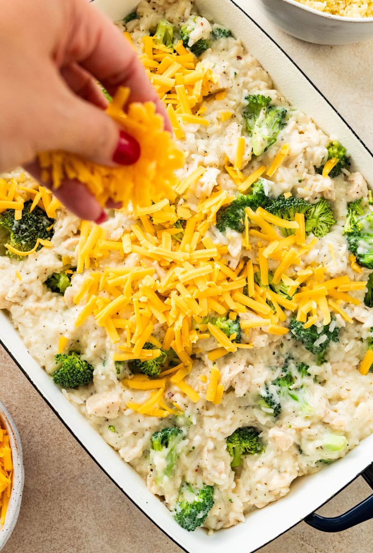baked chicken, broccoli, and rice in a casserole dish ready to bake. Hand sprinkling cheese on the top.
