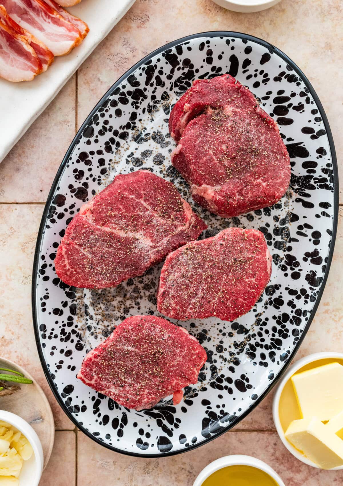 four uncooked filet mignon steaks on a plate after seasoning with salt and pepper.
