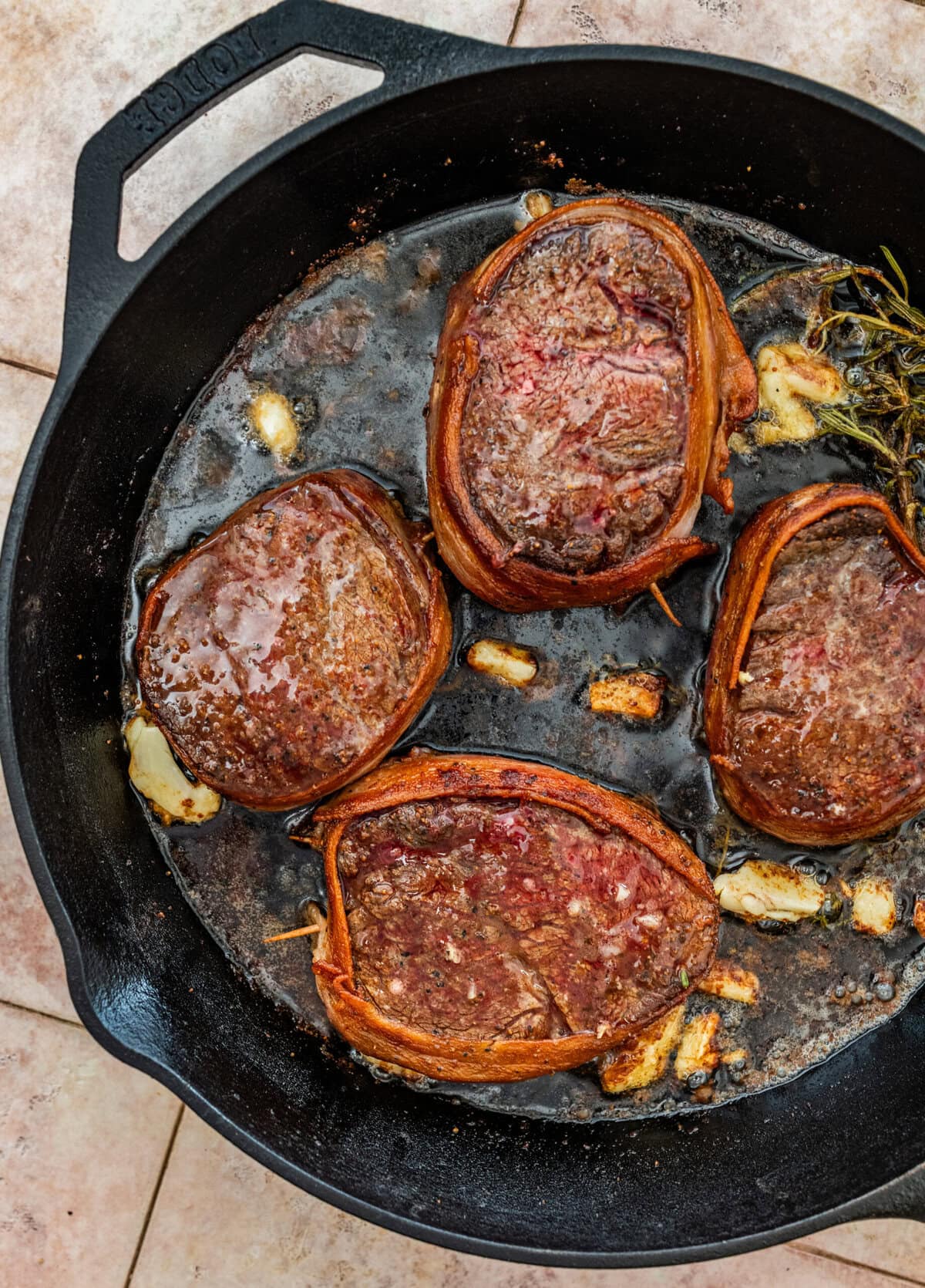 four bacon wrapped filet mignons on a skillet, showing perfectly seared edges and garlic and herbs in pan.
