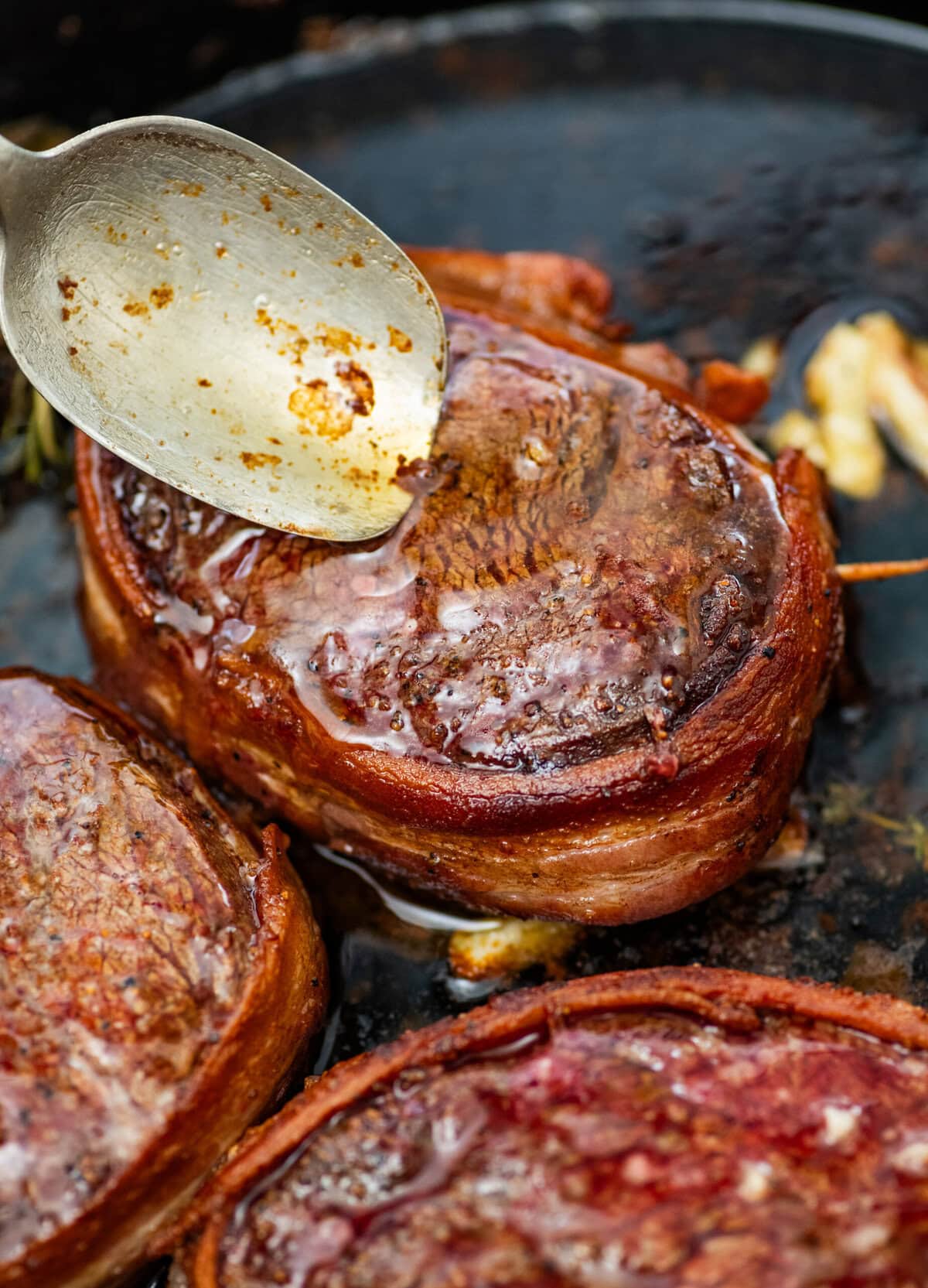 spooning juices over a bacon wrapped filet mignon with other steaks in the background.