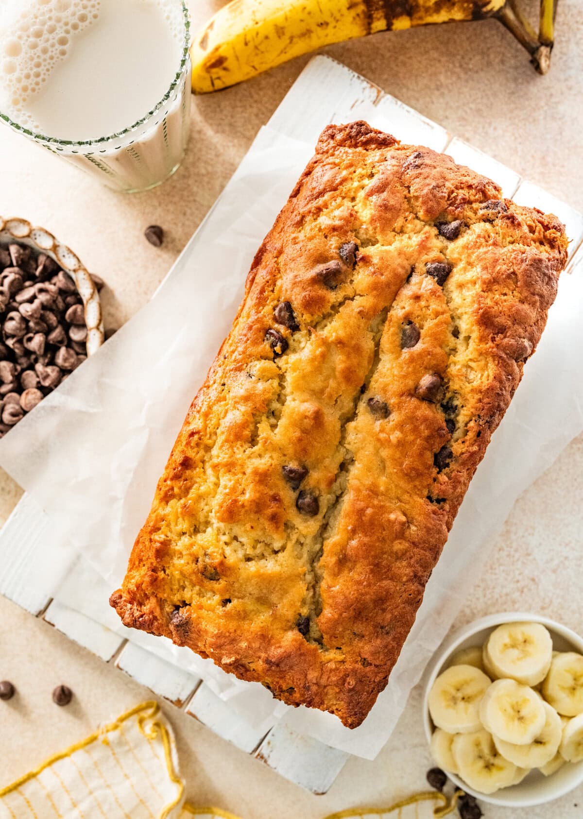 A loaf of chocolate chip banana bread with chocolate chips and bananas in bowls beside it.