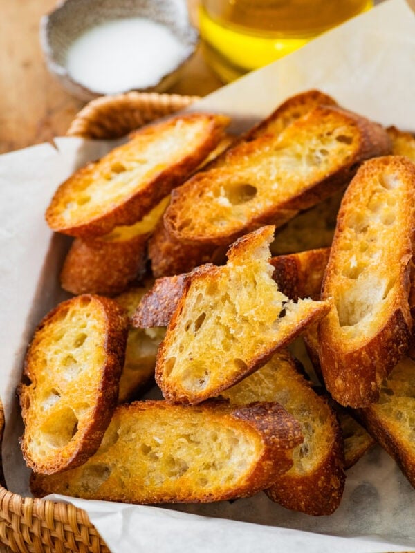 A pile of crostini on a tray with one piece missing a bite and salt and olive oil in the background.