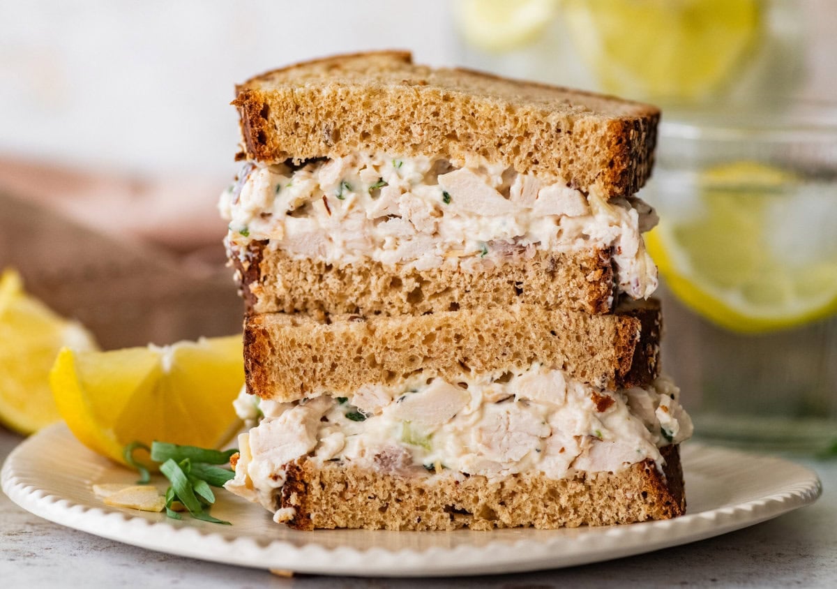 Two halves of a tarragon chicken salad sandwich stacked on top of each other on a plate with lemon.