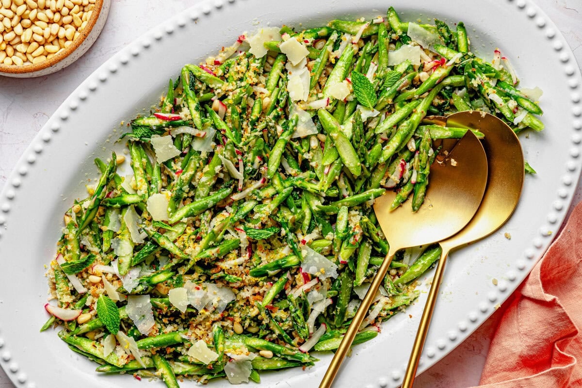 Asparagus salad on a serving dish with serving utensils and extra breadcrumbs in a bowl nearby.