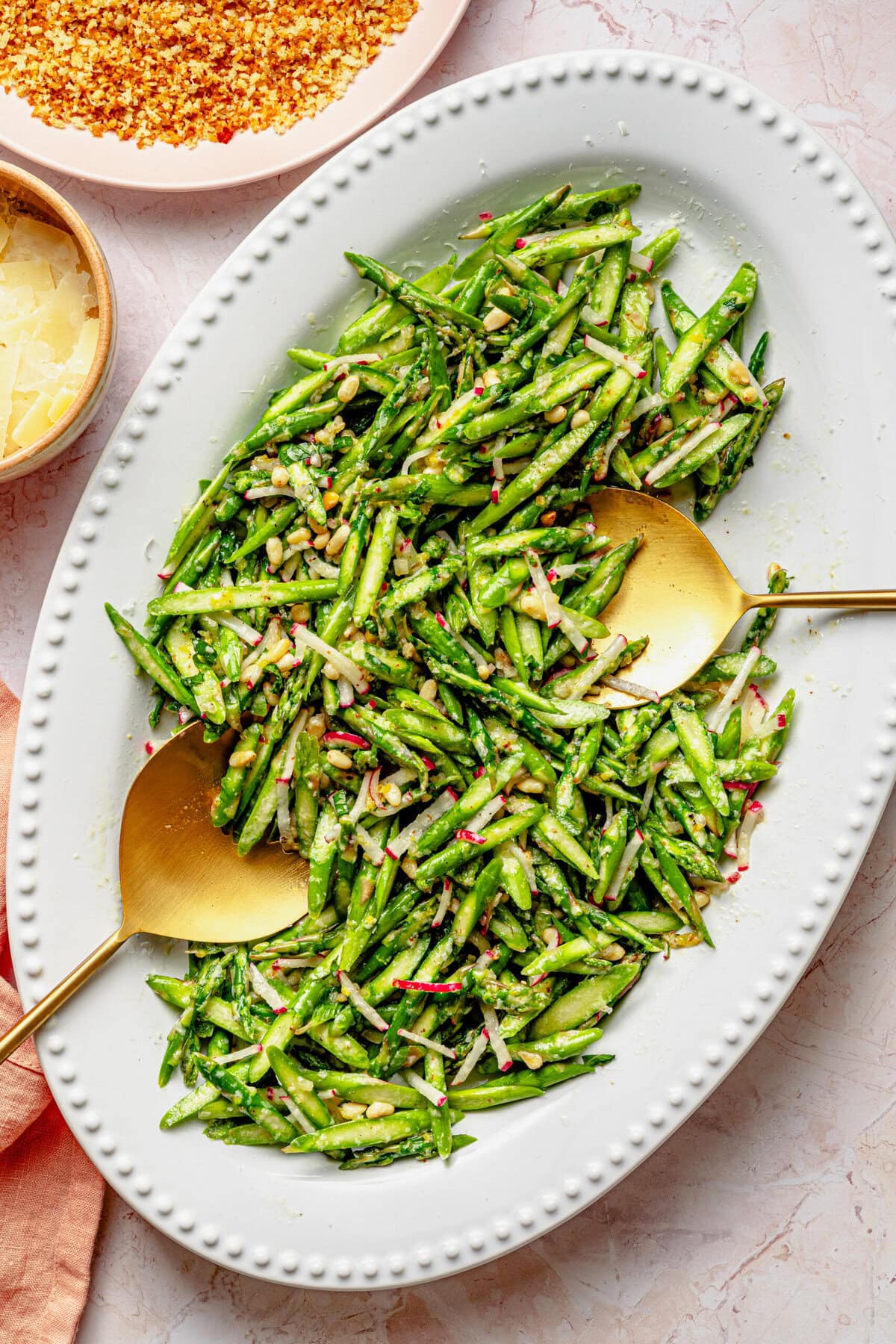 Asparagus salad on a serving dish with serving utensils and extra breadcrumbs in a bowl nearby.