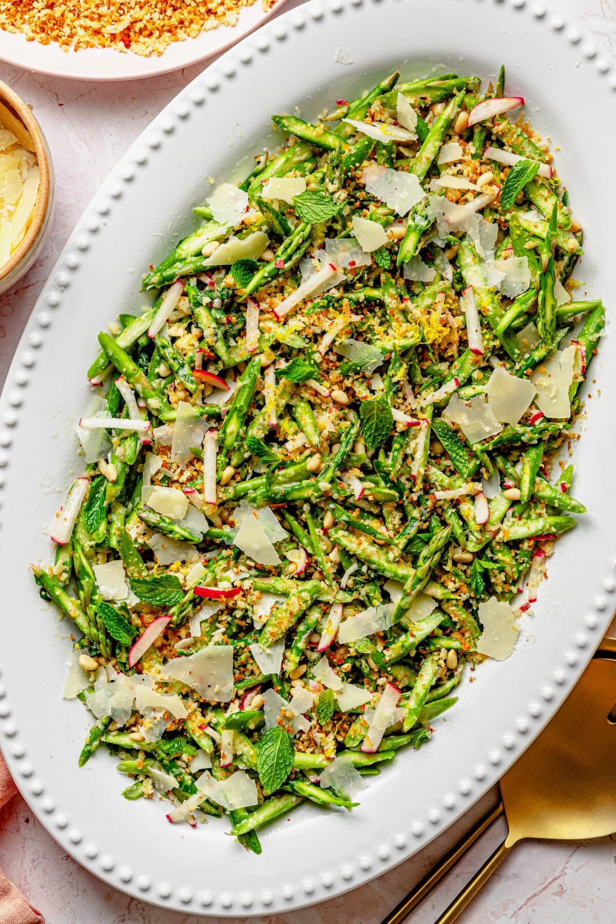 Asparagus salad on a serving dish with serving utensils and extra breadcrumbs in a bowl nearby.