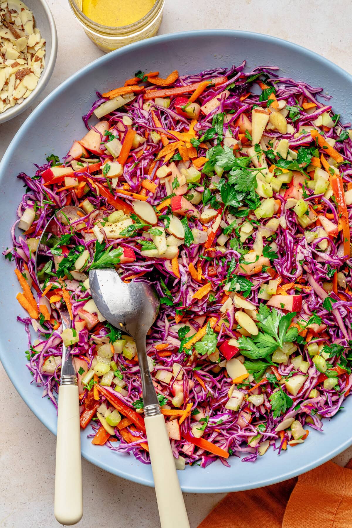 Red cabbage slaw in a bowl with serving utensils, with more ingredients nearby.