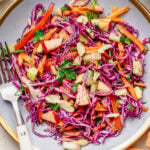 Red cabbage slaw on a plate with a fork with another plate nearby, and extra ingredients.