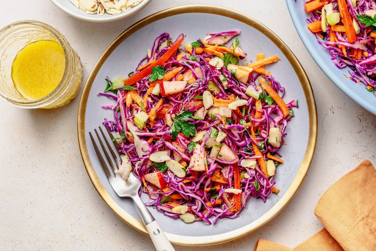 Red cabbage slaw on a plate with a fork with another plate nearby, and extra ingredients.