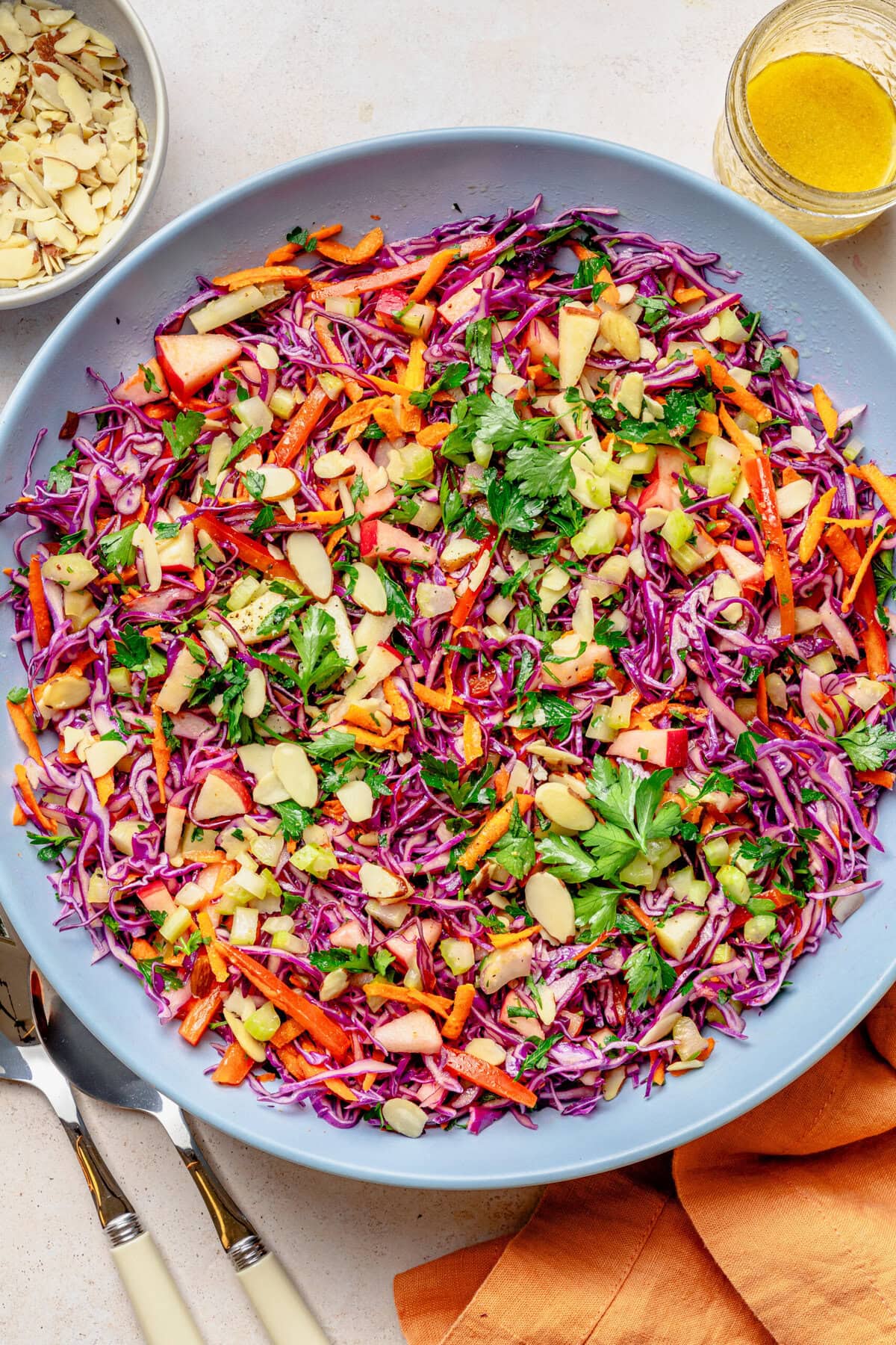 Red cabbage slaw in a bowl with serving utensils, with more ingredients nearby.