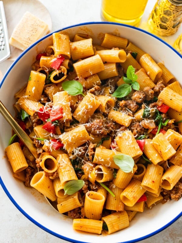 A serving bowl of sausage and peppers pasta topped with parmesan with extra ingredients in the background.