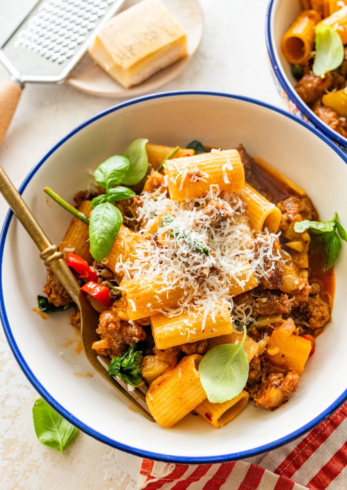 A single portion bowl of sausage and peppers pasta topped with parmesan cheese with a second bowl in the background,