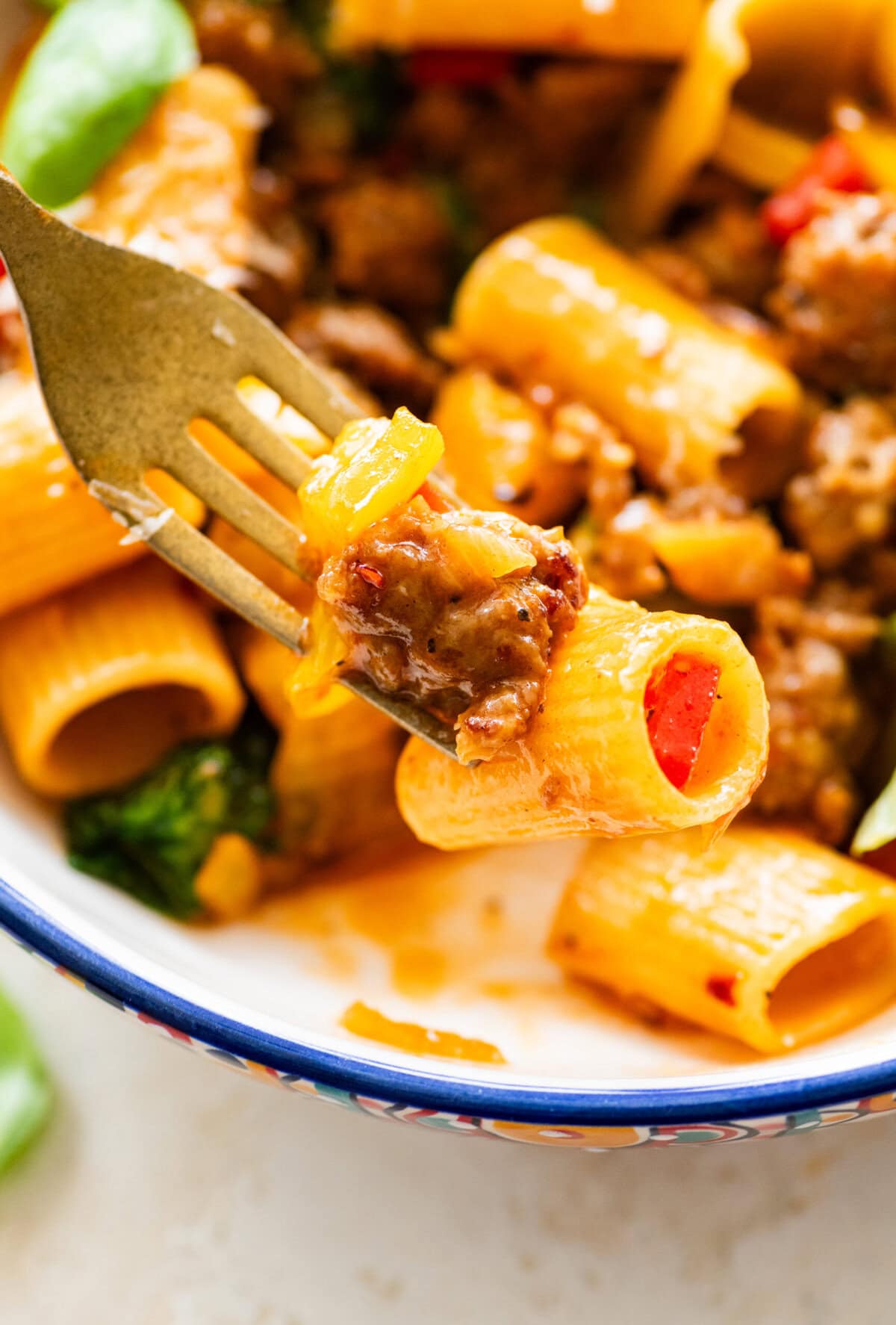 A fork holding up one bite of sausage and peppers pasta over a bowl of more of the recipe.