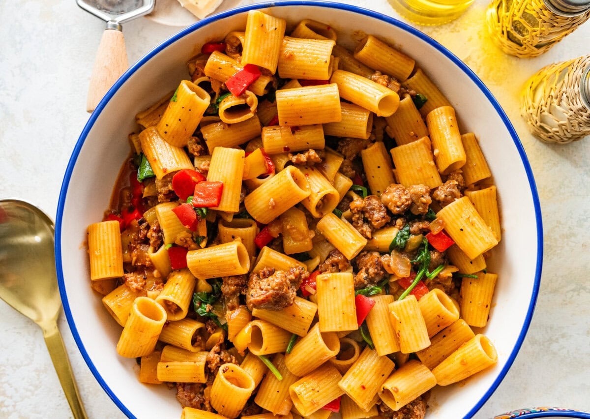 A serving bowl of sausage and peppers pasta with extra ingredients in the background.
