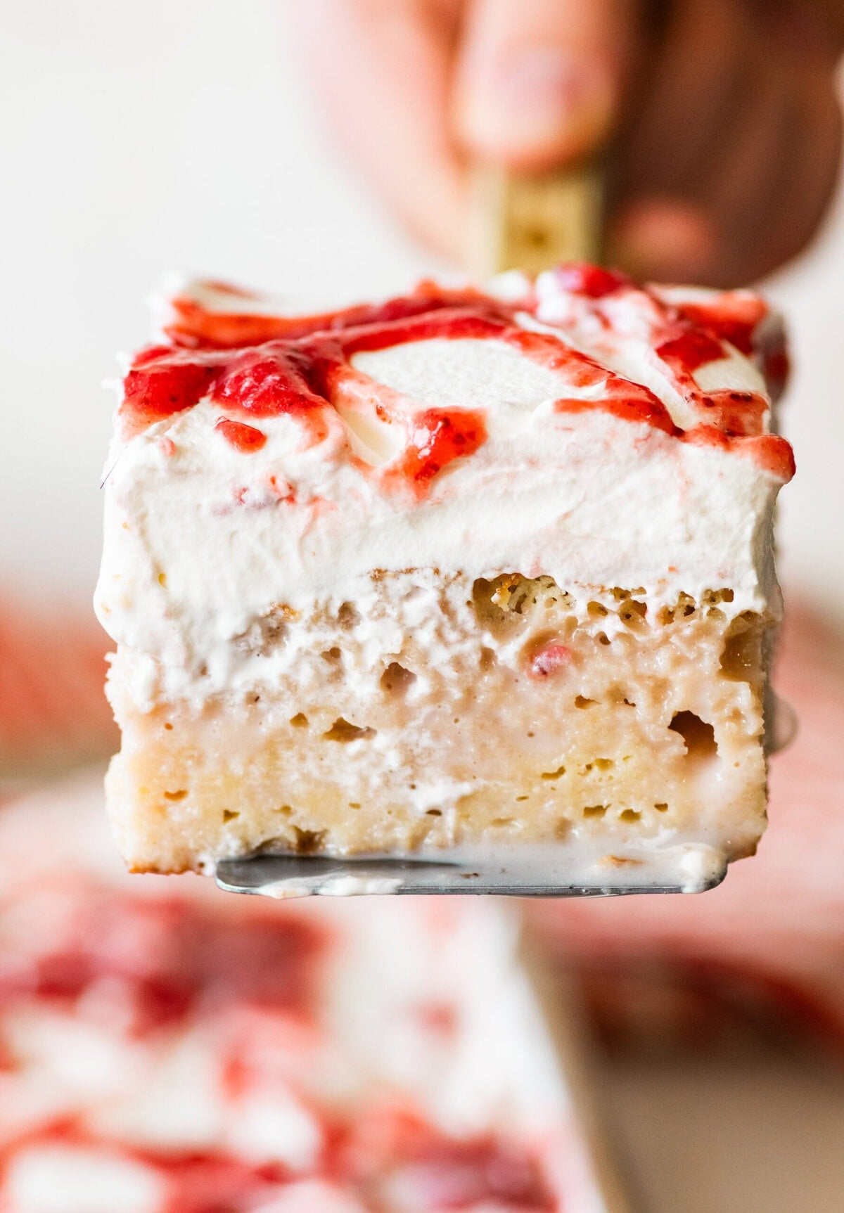 a slice of strawberry tres leches cake shown from the side, being held up by a cake server.