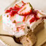 a slice of strawberry tres leches cake with a bite taken out, shown from the side, plated with a spoon and more ingredients nearby.