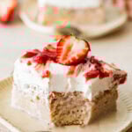 a slice of strawberry tres leches cake with a bite taken out, shown from the side, plated with a spoon and more ingredients nearby.