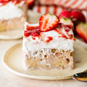 a slice of strawberry tres leches cake, shown from the side, plated with a spoon and more ingredients nearby.