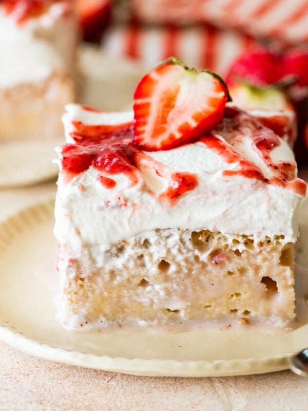 a slice of strawberry tres leches cake, shown from the side, plated with a spoon and more ingredients nearby.
