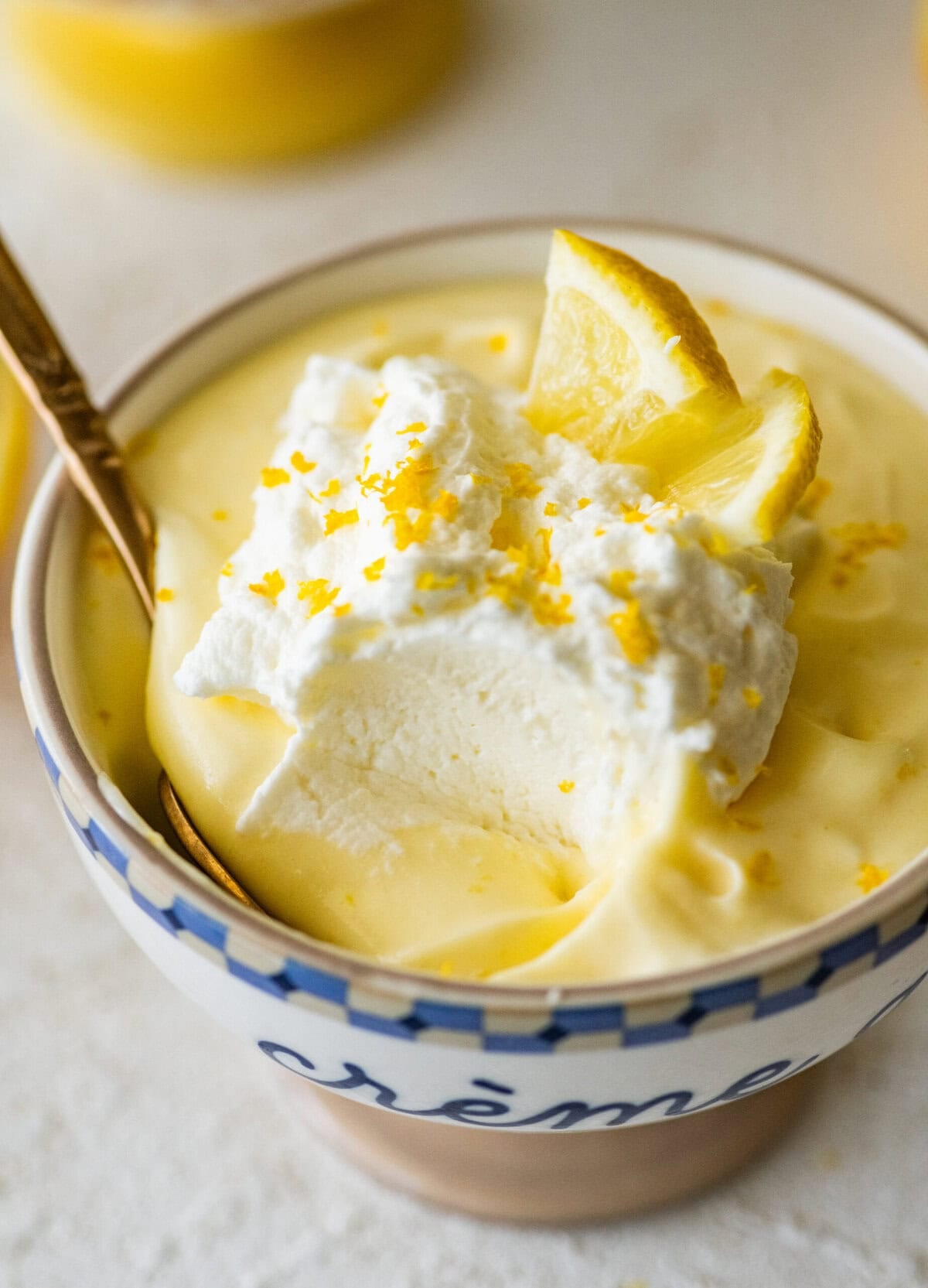 One serving of lemon mousse in a bowl with garnish and more ingredients in the background and a spoon resting inside.