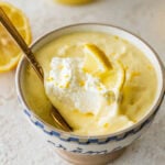 One serving of lemon mousse in a bowl with garnish and more ingredients in the background and a spoon resting inside.