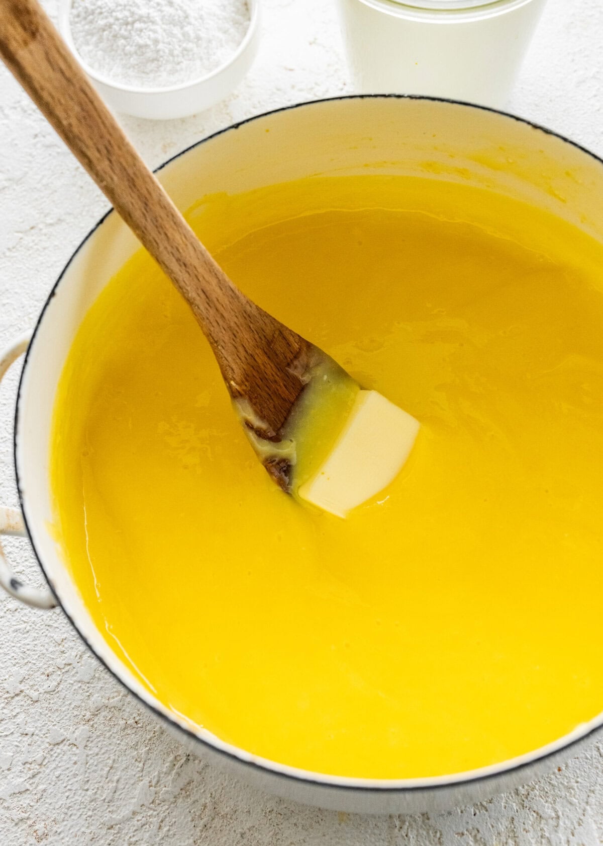 Pats of butter inside the pot of lemon curd with a wooden spoon mixing it around.