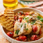 A plate of italian poached fish with garnish, bread, and more ingredients nearby.