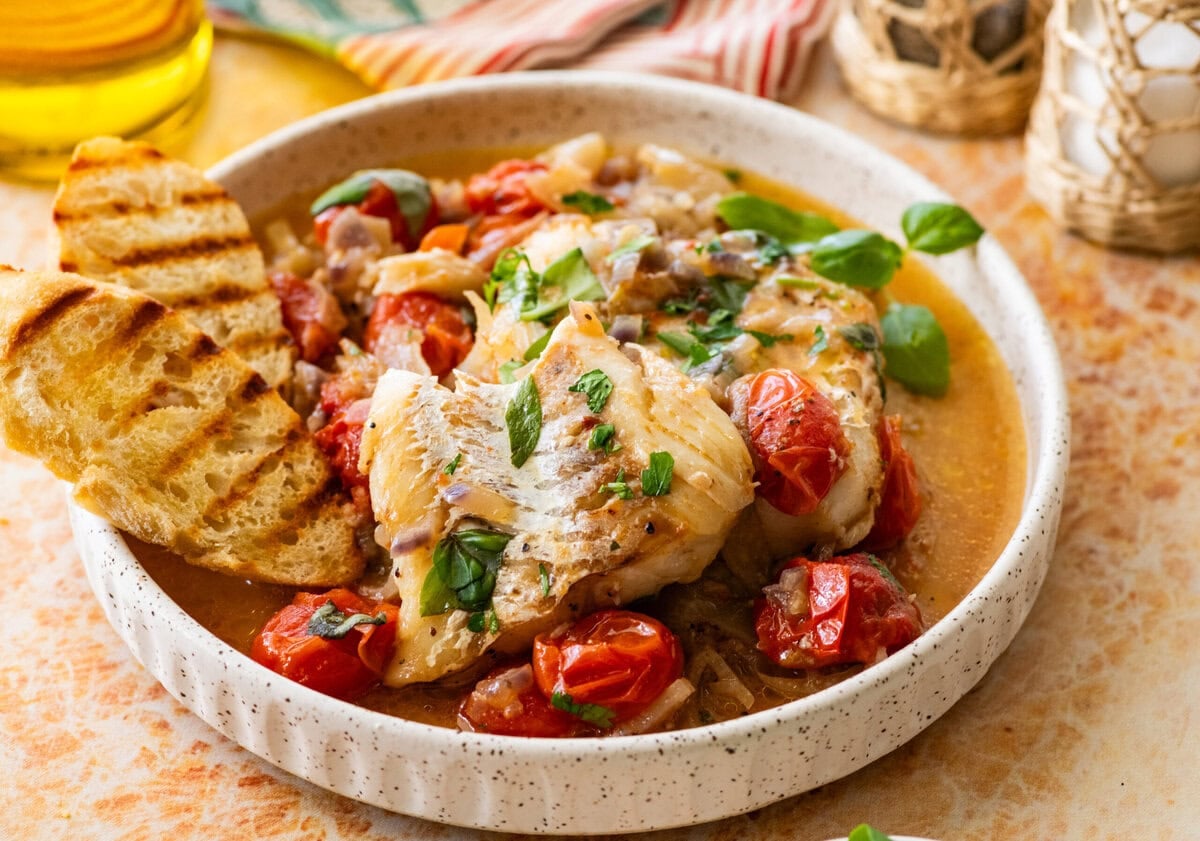 A plate of italian poached fish with garnish, bread, and more ingredients nearby.