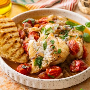 A plate of italian poached fish with garnish, bread, and more ingredients nearby.