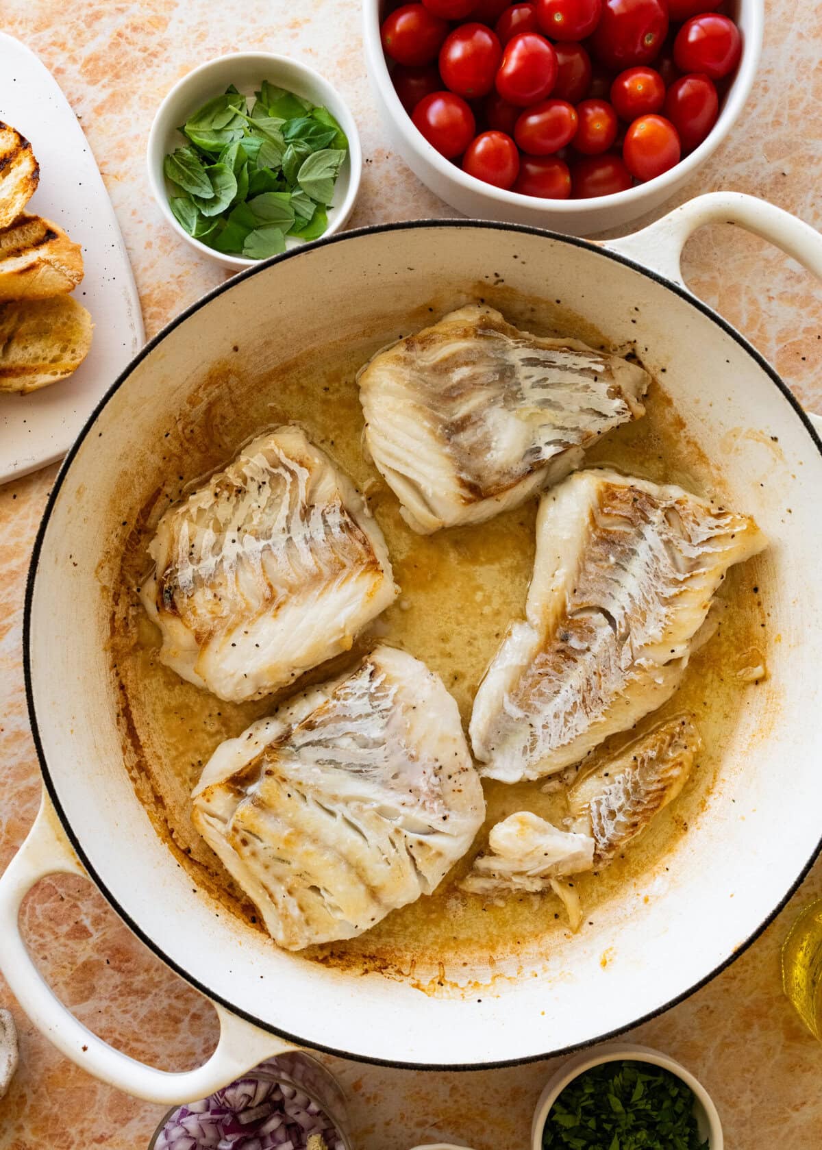 Seasoned fish in a skillet with oil and more ingredients nearby.