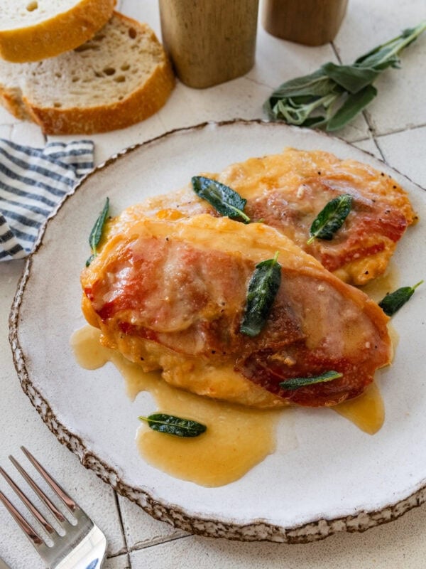 two pieces of chicken saltimbocca on a plate, garnished with sage, with more ingredients nearby.