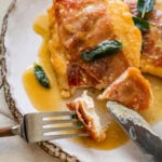 a fork and knife cutting into a piece of chicken saltimbocca that is plated and garnished with sage.