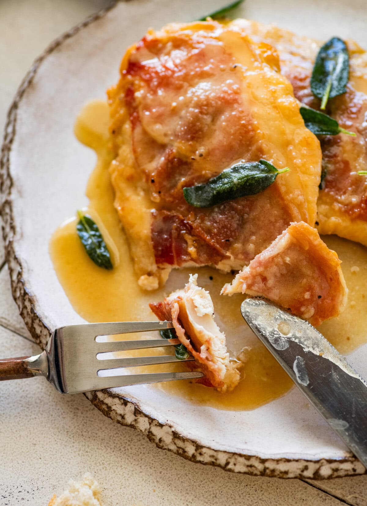 a fork and knife cutting into a piece of chicken saltimbocca that is plated and garnished with sage.