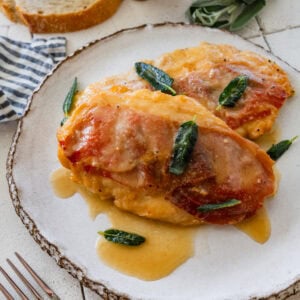 two pieces of chicken saltimbocca on a plate, garnished with sage, with more ingredients nearby.