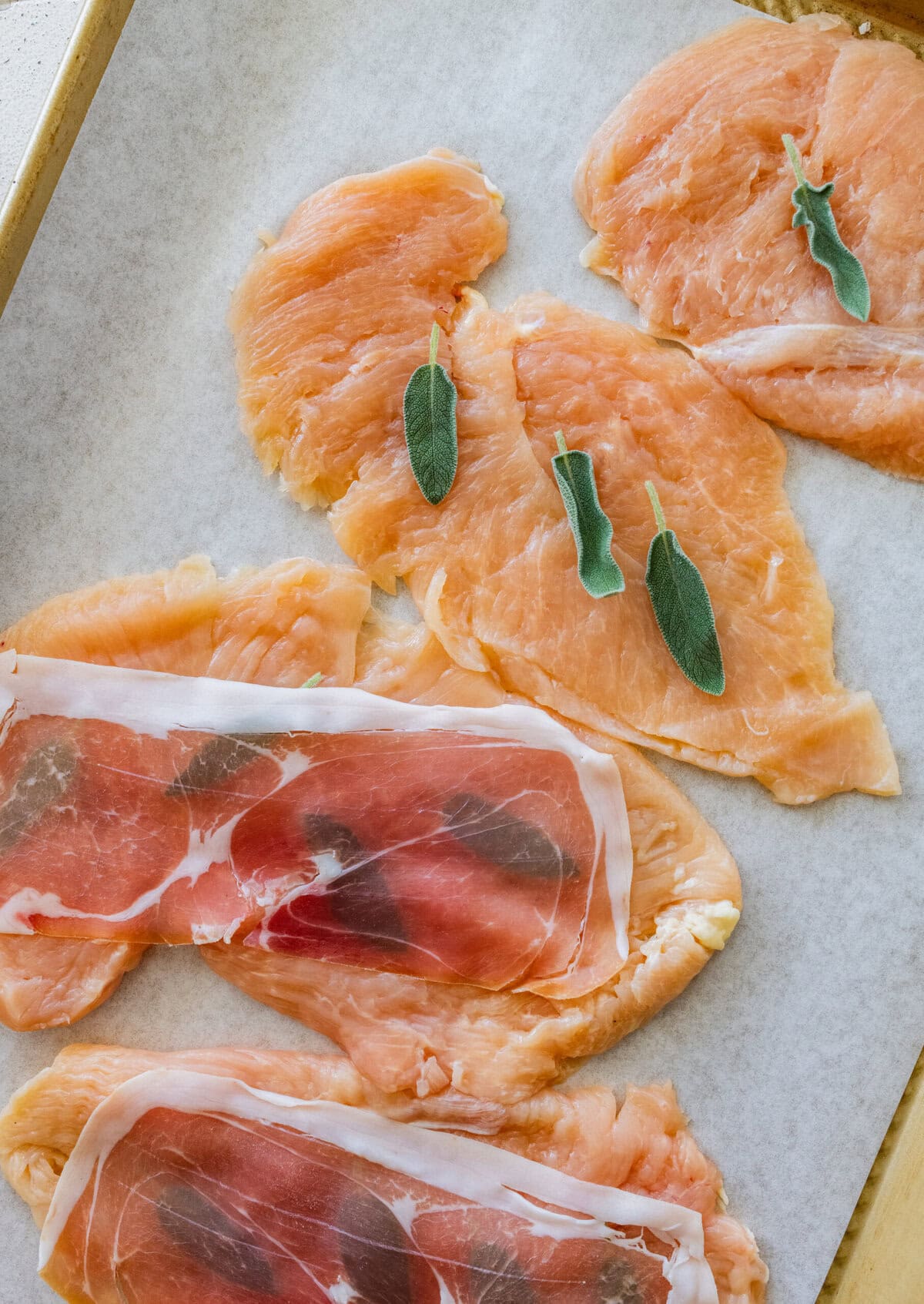 thin, pounded chicken breasts on a tray with sage leaves on top, with prosciutto being placed on top.