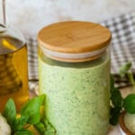 a clear jar of creamy pesto sauce with more ingredients nearby.
