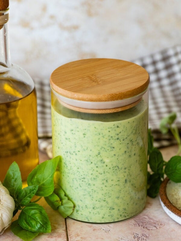 a clear jar of creamy pesto sauce with more ingredients nearby.