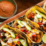 3 fish tacos with toppings, placed on a tray with more ingredients to make the recipe nearby.