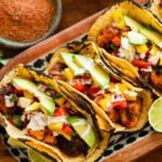 3 fish tacos with toppings, placed on a tray with more ingredients to make the recipe nearby.