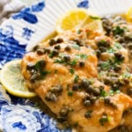 Two breasts of lemon caper chicken on a dish garnished with lemon slices, and more ingredients nearby.