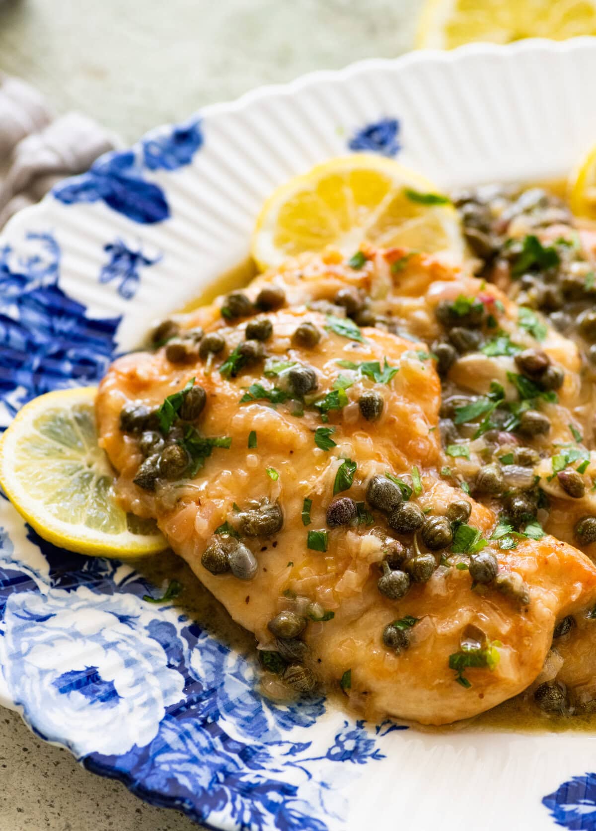Two breasts of lemon caper chicken on a dish garnished with lemon slices, and more ingredients nearby.