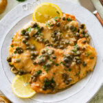Two breasts of lemon caper chicken on a dish garnished with lemon slices, and more ingredients and utensils nearby.