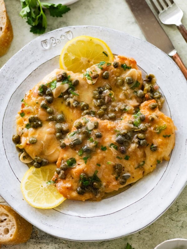 Two breasts of lemon caper chicken on a dish garnished with lemon slices, and more ingredients and utensils nearby.